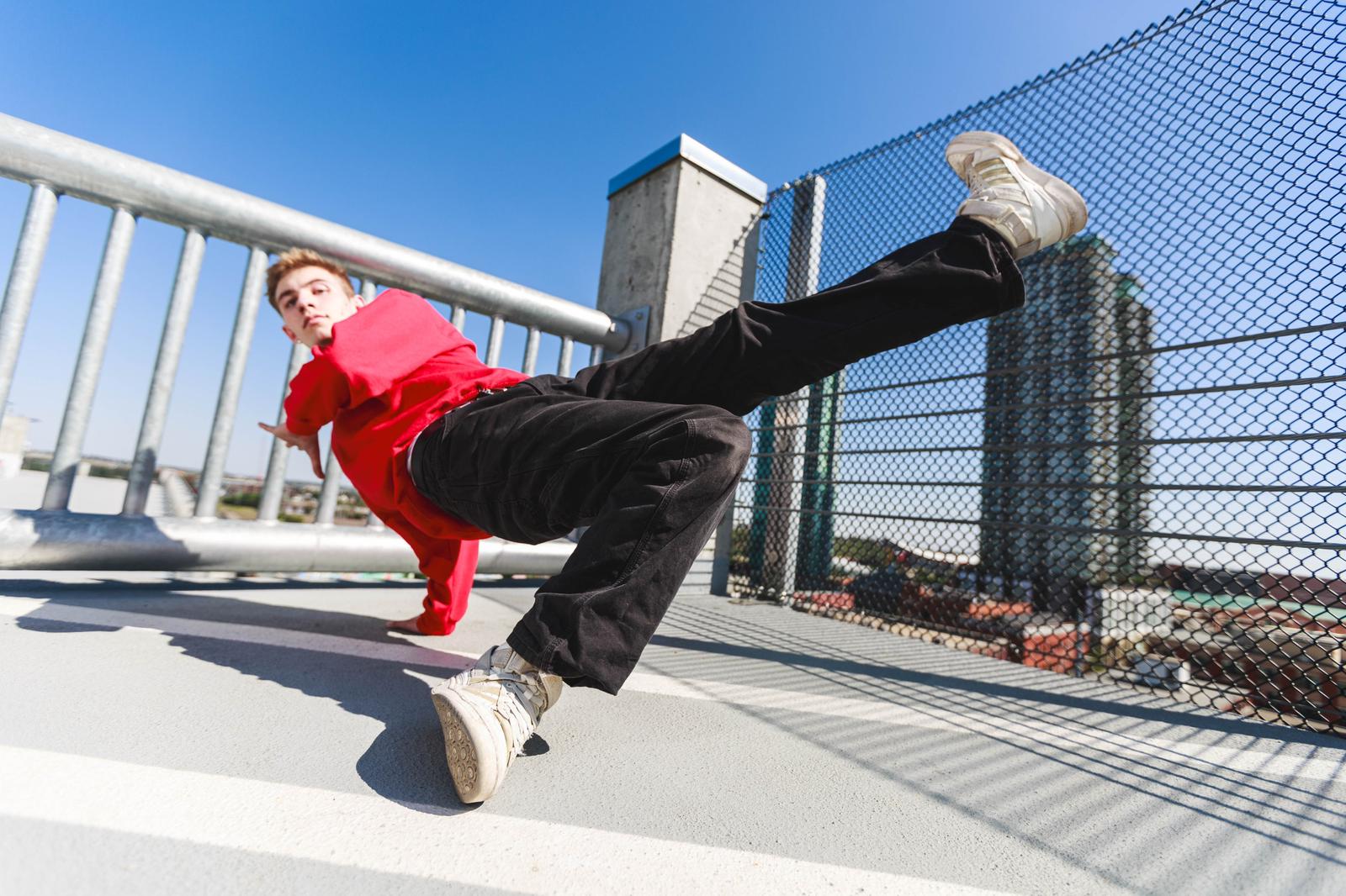 Eli Everett, PULSE’s newest MVP, performs a dynamic Breaking move on a rooftop with a cityscape in the background. Wearing a red sweatshirt and black pants, he executes a powerful freeze, emphasizing his skill and passion for Breaking.