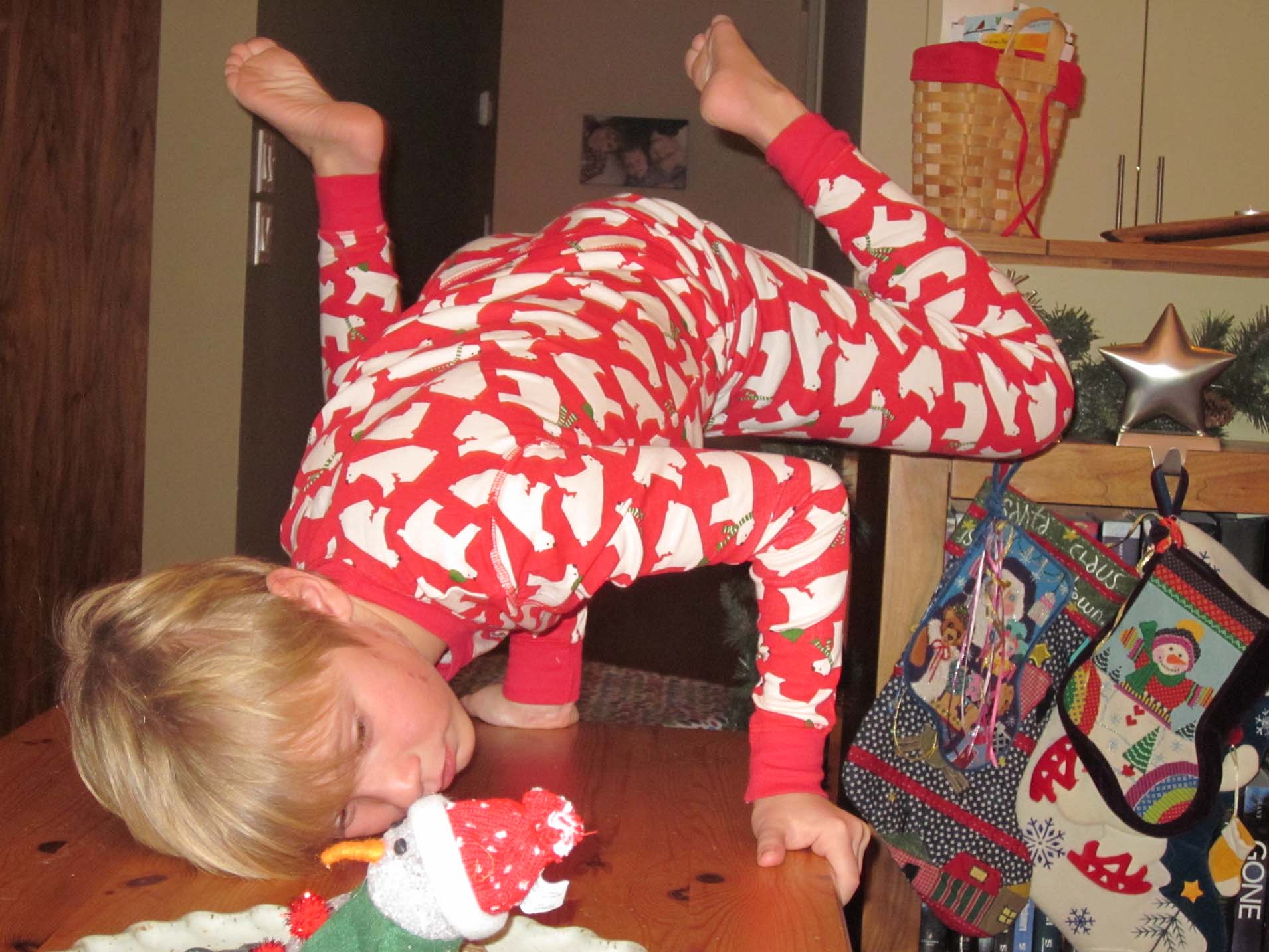 A young Eli in festive red pyjamas with white polar bears performs a B-Boy pose on a wooden table during the holiday season. The background features Christmas stockings and decorations, capturing an early moment in his dance journey.