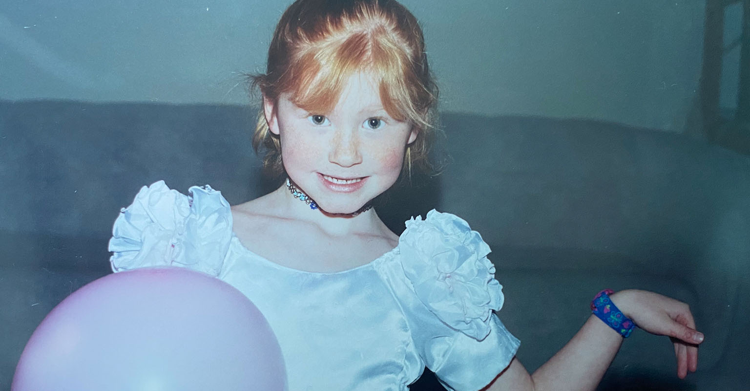 Childhood photo of Beniya Lawrence smiling in a white tap dance costume while holding a pink balloon.